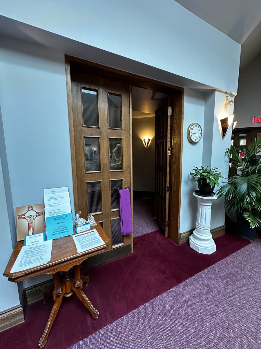 Narthex hallway showing the confessional/reconciliation room entrance with purple stole hung on door, an examination-of-conscience literature table, wall clock, exit sign, and potted plants on burgundy carpet Clear, in-focus, informative but visually busy (exit sign, wall clock, pedestal plant, paper handouts) — reads more administrative than serene. Useful documentary shot of the reconciliation room location; not a strong hero image. Slight wide-angle distortion from close framing.