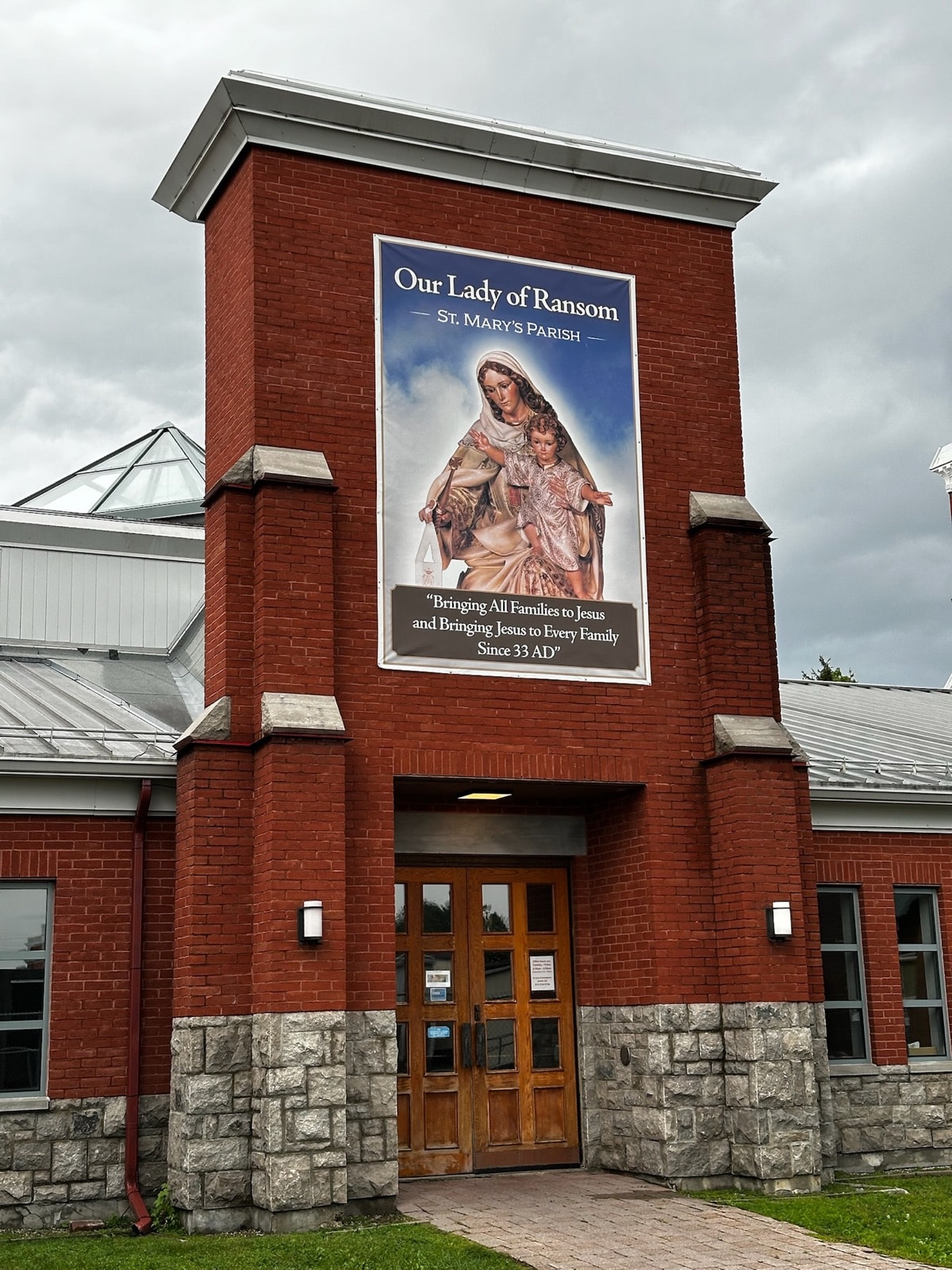 Red-brick entrance tower with a large banner of Our Lady of Ransom holding the Child Jesus, above wooden double doors reached by a level brick walkway.