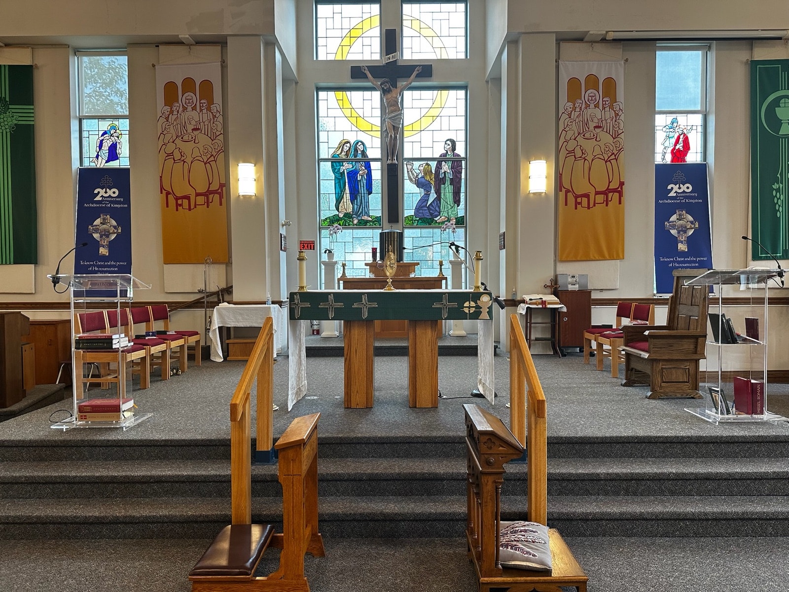 The sanctuary seen head-on: a stone altar dressed in a green cloth, flanked by reading stands, beneath a crucifix and a large stained-glass window of the Crucifixion.