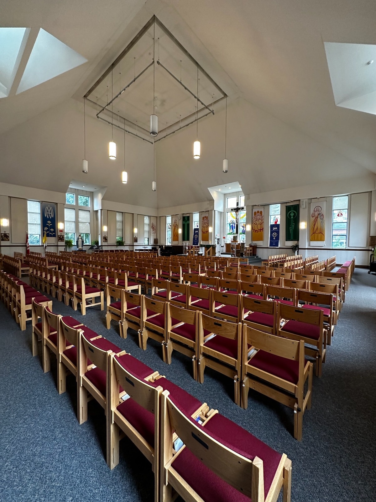 Wide view across rows of light wooden chairs with burgundy cushions, beneath a white vaulted ceiling with skylights and hanging pendant lights.