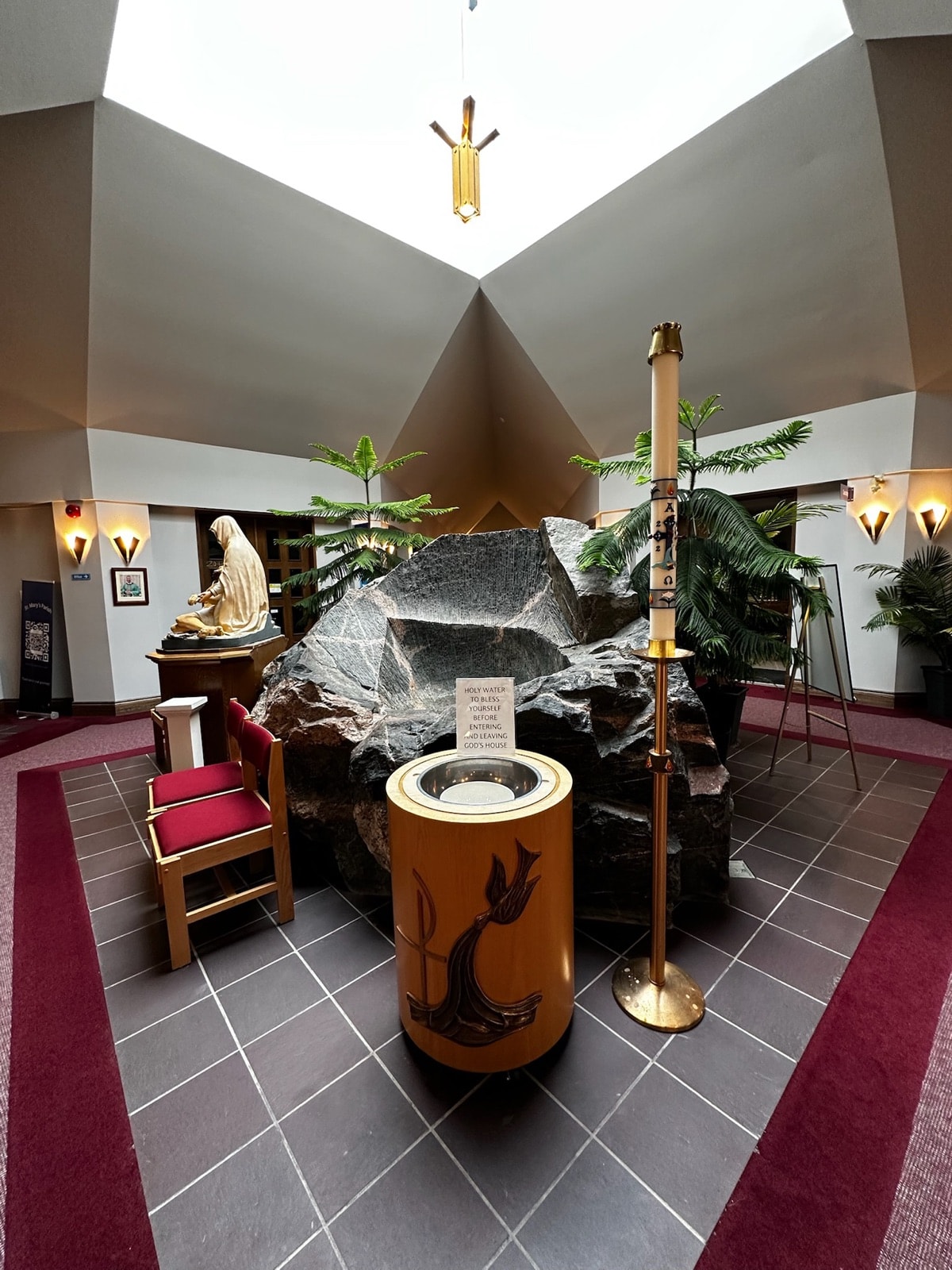 A large natural rock beneath a skylight, with a carved wooden baptismal font bearing a dove and Chi-Rho, a tall paschal candle, and a statue to one side.